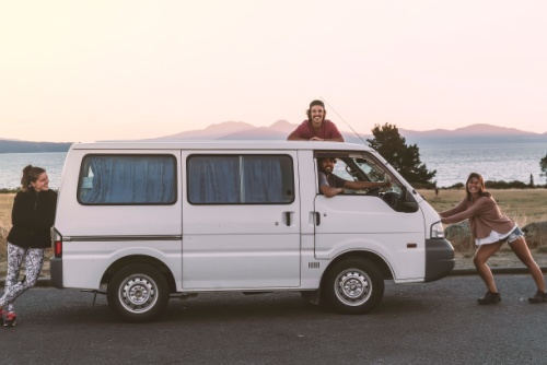 A group of 4 happy friends (2 girls and 2 boys) who love traveling stay close to their van at sunset. They travel with this simple and cheap white camper van as backpackers on a budget. Carpe diem