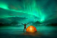 A man camping in wild northern mountains with an illuminated tent viewing a spectacular green northern lights aurora display. Photo composition. A man camping in wild northern mountains with an illuminated tent viewing a spectacular green northern lights aurora display. Photo composition.
